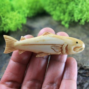 Puede incluir: Una figurita de pescado de hueso tallada a mano. El pez es de color marrón claro y tiene tallas detalladas de escamas y aletas. El pez mide aproximadamente 10 cm de largo.