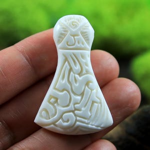 May include: A cream-colored, carved pendant in the shape of an axe. The pendant features intricate, swirling patterns and a detailed design on the head of the axe. The background is blurred, highlighting the pendant's craftsmanship.