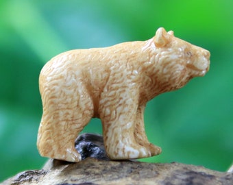 May include: A small, tan-colored bear figurine with detailed fur texture. The bear is standing on all fours, with its head turned slightly to the side. The figurine is made of a light-colored material and is set against a blurred green background.
