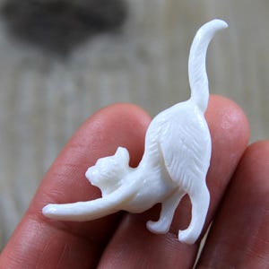 May include: A small, white cat figurine in a stretching pose. The cat is sculpted with detailed fur texture and has its tail arched upwards. The figurine is held in a person's hand, showcasing its size and design.