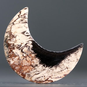 May include: A crescent-shaped decorative object with a polished surface. The object features a striking contrast of colors and textures, with areas of black, cream, and brown. The overall shape resembles a stylized moon, suitable for jewelry or decorative purposes.
