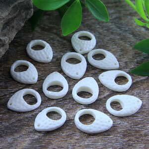 May include: A collection of white, oval-shaped, shell pendants with a textured surface and a hole for stringing. Each pendant has a smooth, circular opening in the center.
