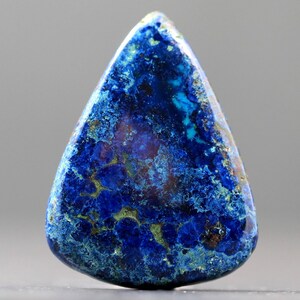 May include: A polished, teardrop-shaped gemstone with a deep blue hue, accented by lighter blue and green veins. The stone's surface has a textured appearance, with areas of darker and lighter coloration. The overall effect is a striking display of natural beauty.