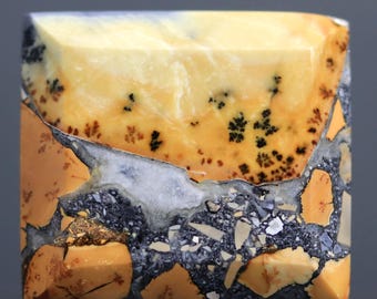 May include: A square-shaped gemstone with a mix of colors. The top section is a light yellow with black speckles, transitioning to a mix of orange, gray, and white. The bottom section has a mosaic-like appearance with orange and gray fragments.