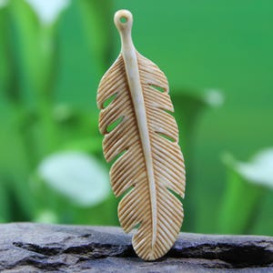 May include: A carved bone feather pendant with a detailed design. The feather is light brown in color and has a hole at the top for hanging.