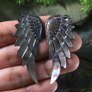 May include: Two dark gray, iridescent wing-shaped pendants. Each wing has a detailed feather design with a pearlescent sheen. The wings are likely jewelry components, possibly earrings or pendants, with a dark, almost black, color.