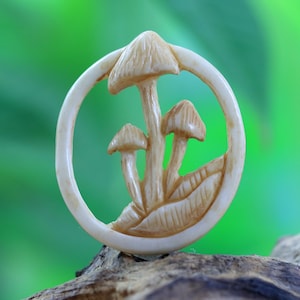 May include: A white carved wooden pendant featuring three mushrooms in a circle. The mushrooms are carved in detail and have a realistic appearance.