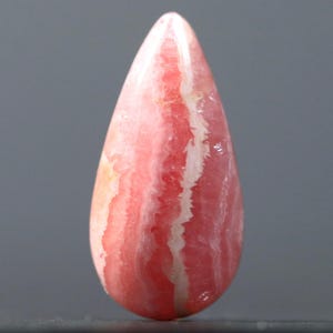 May include: A teardrop-shaped pink and white banded gemstone cabochon. The stone has a smooth, polished surface and is translucent.