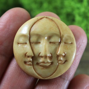 以下が含まれることがあります： 三日月形に彫刻された木製のペンダント。3つの顔があり、目は閉じ、口は微笑んでいます。ペンダントは薄いベージュ色です。