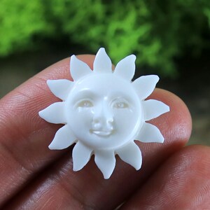 Könnte beinhalten: Eine weiße, geschnitzte Muschelsonne mit einem lächelnden Gesicht. Die Sonne hat ein detailliertes Gesicht mit Augen, einer Nase und einem Mund.