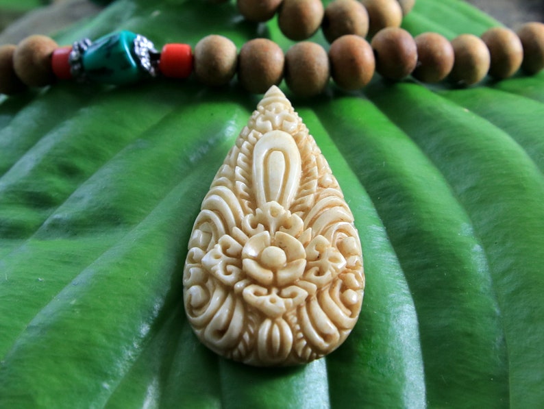 Puede incluir: Un colgante blanco tallado con intrincados dise&ntilde;os florales, que se asemeja a una l&aacute;grima. El colgante est&aacute; apoyado sobre una hoja verde.