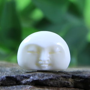 May include: A small, white, carved figurine of a face with closed eyes and a smiling mouth. The figurine is made of bone or ivory and is a traditional Japanese charm.