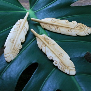 May include: Three carved bone feathers with intricate designs. The feathers are white and have a natural bone color. They are arranged on a green leaf.