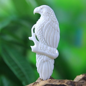 May include: Ivory-colored carved eagle figurine perched on a branch. The eagle is detailed with textured feathers and a sharp beak. The figurine is set against a blurred green background, highlighting the intricate carving.