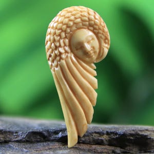 May include: A hand-carved ivory pendant featuring a stylized human face with a feathered headdress. The pendant is shaped like a wing and has a smooth, cream-colored finish.