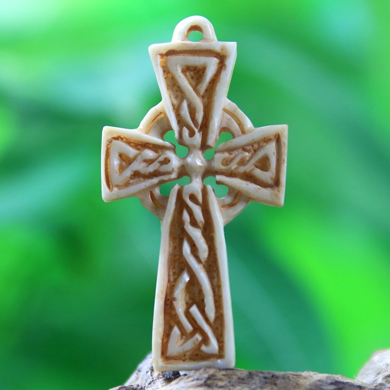 May include: A white Celtic cross pendant with intricate knotwork design. The cross is carved from bone and has a light brown patina.