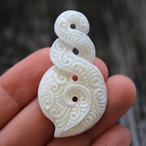 May include: White bone pendant with intricate carvings in a traditional Maori design. The pendant has two holes for stringing.