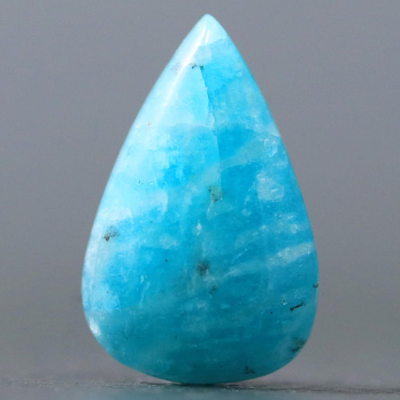 May include: A teardrop-shaped cabochon of blue amazonite gemstone. The stone has a light blue color with white and gray streaks throughout.