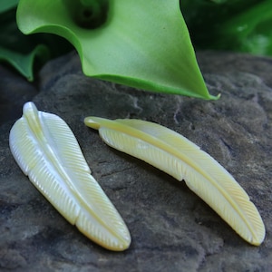 May include: Two white mother of pearl feather charms with a detailed carved design. The feathers are laying on a dark brown textured surface.