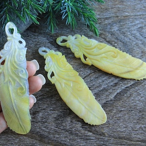 Peut inclure: Trois pendentifs en forme de plume en coquille sculptée, de couleur jaune et blanche. Les plumes présentent des motifs complexes et sont d'une couleur jaune clair avec des accents blancs.