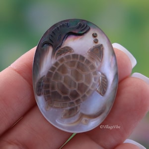 Puede incluir: Un colgante de nácar tallado con una tortuga con un diseño de caparazón detallado. La tortuga está rodeada de burbujas y una ola estilizada.