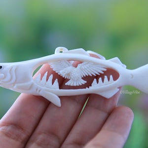 May include: White bone carving of a salmon with a bird flying out of its body. The carving is detailed and intricate, with the bird's wings spread out and the salmon's scales visible.