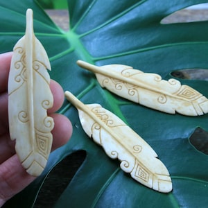 May include: Three carved bone feathers with intricate designs. The feathers are light brown and have a smooth, polished finish.