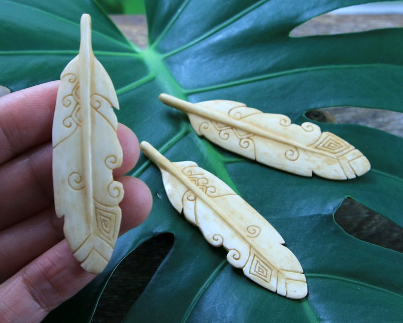May include: Three carved bone feathers with intricate designs. The feathers are light brown and have a smooth, polished finish.