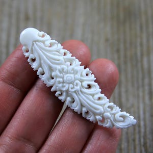以下が含まれることがあります： 象牙色の彫刻ペンダント。複雑な花柄と渦巻き模様のデザイン。細長い形状で、丸みを帯びた上部と詳細な対称パターンが特徴です。この装飾品は、ジュエリーやクラフトプロジェクトに使用される可能性があります。