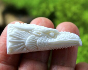 May include: A carved, off-white eagle head sculpture. The sculpture is highly detailed, with visible feathers, an open beak, and a focused eye. The eagle head is held in a person's hand, with a blurred green background.