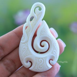 Puede incluir: Pendiente de hueso blanco tallado con un diseño tradicional maorí, que presenta una espiral y patrones intrincados. El pendiente tiene forma de gancho.