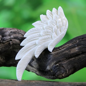 May include: A white carved bone wing with detailed feather patterns. The wing is resting on a piece of weathered wood.