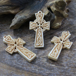 May include: Three intricately carved Celtic cross pendants made of white bone. The crosses feature a knotwork design and are displayed on a wooden surface.