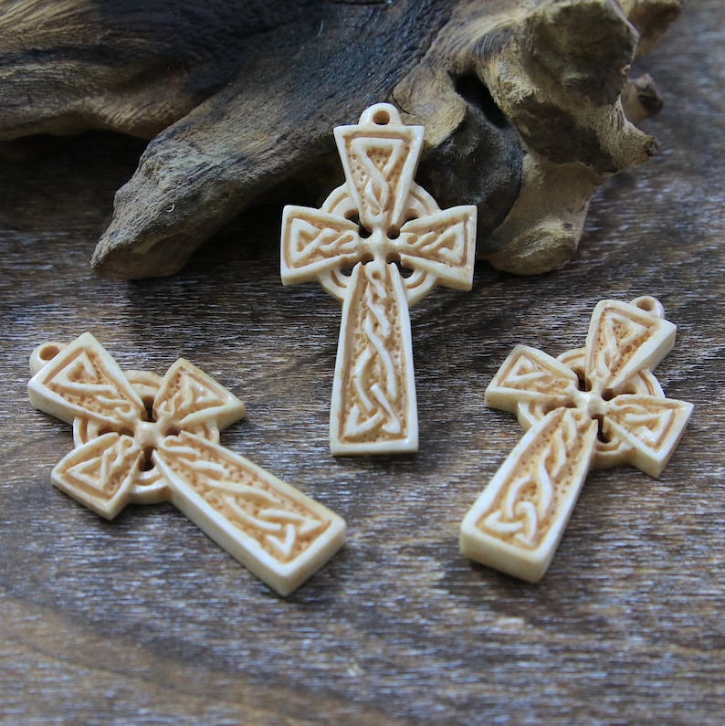 May include: Three intricately carved Celtic cross pendants made of white bone. The crosses feature a knotwork design and are displayed on a wooden surface.