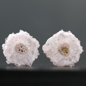 May include: Two white quartz crystal clusters with a central, circular pattern. The centers have a mix of brown, green, and tan hues. The crystals have a spiky, textured appearance, and are set against a gray background.