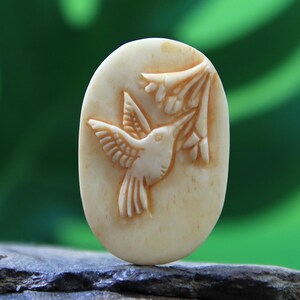 May include: An oval bone pendant carved with a detailed hummingbird design. The hummingbird is shown with its beak in a flower.