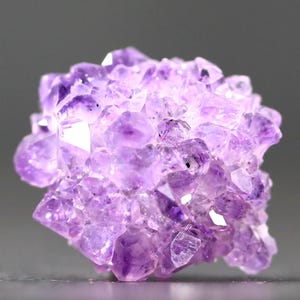 May include: A cluster of amethyst crystals, showcasing a range of purple hues from light lavender to deep violet. The crystals are sharply pointed and densely packed, creating a textured, irregular shape. The gemstone is set against a neutral gray background.