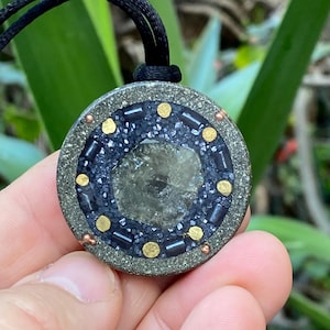 Op de afbeelding: Ronde hangerketting met een centrale, lichtgekleurde steen omgeven door een donkerblauw en grijs mozaïekontwerp. Het mozaïek is geaccentueerd met goudkleurige stippen en zwarte rechthoekige kralen. De hanger is bevestigd aan een zwart koord.