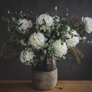 May include: A bouquet of white flowers arranged in a grey and brown ceramic vase. The flowers are surrounded by green leaves.