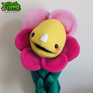 May include: A bright yellow felt flower puppet with pink petals and a green stem. The puppet has a smiling face with black eyes, a white nose, and a black mouth.