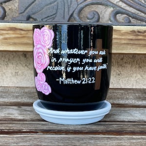 May include: Black ceramic planter with a white saucer. The planter has a pink rose design and the text "And whatever you ask in prayer, you will receive, if you have faith - Matthew 21:22".
