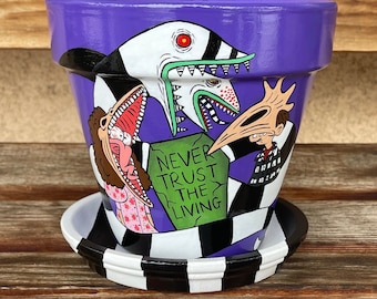 May include: Purple ceramic planter with a black and white checkerboard design. The planter features a hand-painted illustration of characters from the movie Beetlejuice, including the title character, Lydia Deetz, and the Maitlands. The planter also includes the text "Never Trust the Living".