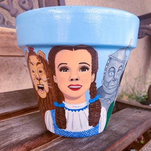 May include: A blue ceramic flower pot painted with characters from the Wizard of Oz. Dorothy is in the center, with the Cowardly Lion on the left and the Tin Man on the right.