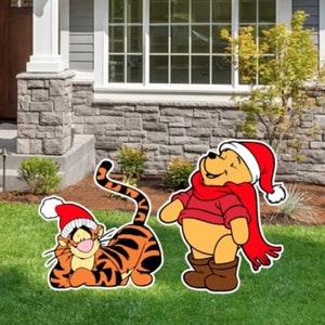 May include: A Winnie the Pooh and Tigger Christmas yard decoration set. Winnie the Pooh is wearing a red scarf and a Santa hat. Tigger is wearing a Santa hat and is lying down. The decorations are in front of a house with a green lawn.