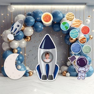 May include: A space-themed party decoration set featuring a large rocket cutout with a child's photo, a crescent moon, and planet cutouts. The backdrop includes a balloon arch in blue, white, and gold, with a cartoon astronaut.