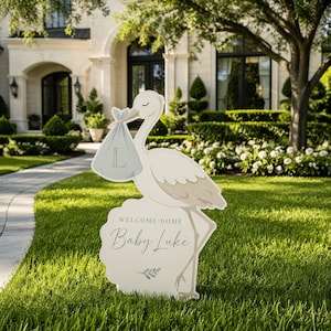 May include: A white stork-shaped lawn sign with a gray baby bundle in its beak. The sign reads "WELCOME HOME Baby Luke" in script. The stork stands on a decorative base with a floral design. The sign is placed on a green lawn.