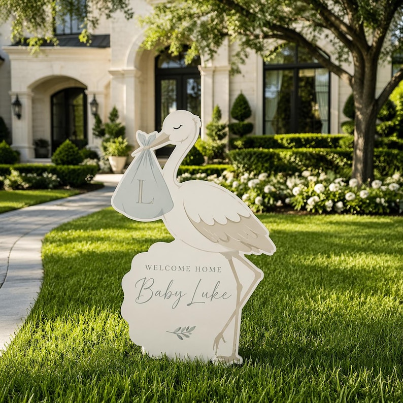 May include: A white stork-shaped lawn sign with a gray baby bundle in its beak. The sign reads "WELCOME HOME Baby Luke" in script. The stork stands on a decorative base with a floral design. The sign is placed on a green lawn.