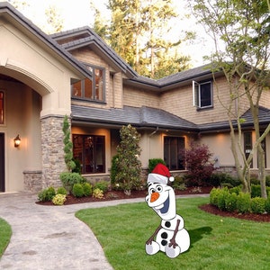 May include: A white snowman wearing a red Santa hat sits on a green lawn in front of a large house with a stone facade.