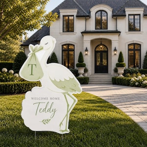 May include: A white and green stork-shaped lawn sign with the text "WELCOME HOME Teddy" in a script font. The stork carries a bundle with the letter "T". The sign is placed on a green lawn in front of a house.