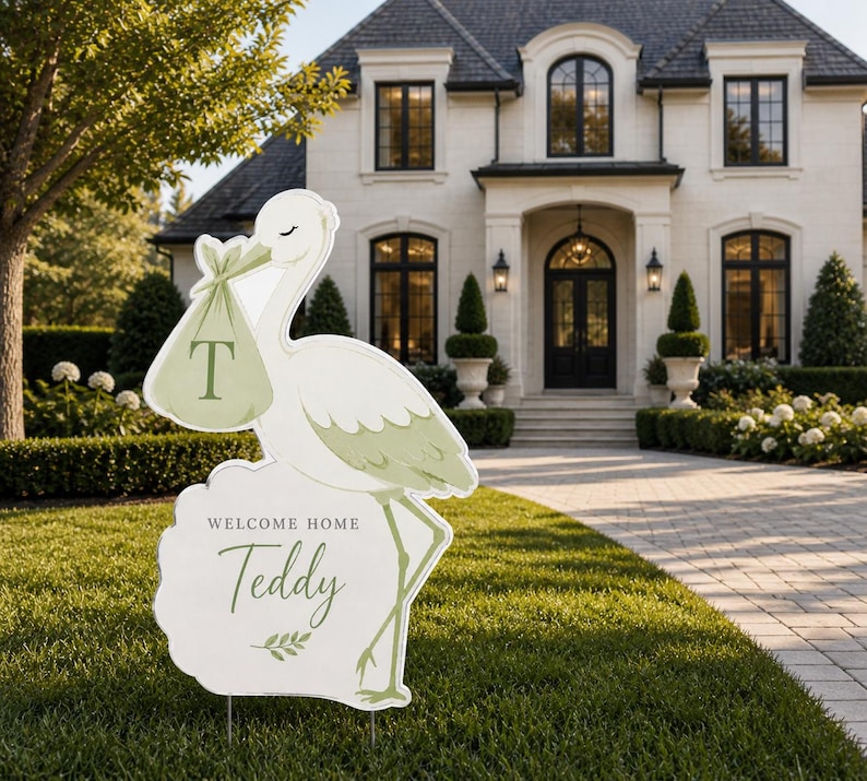 May include: A white and green stork-shaped lawn sign with the text "WELCOME HOME Teddy" in a script font. The stork carries a bundle with the letter "T". The sign is placed on a green lawn in front of a house.